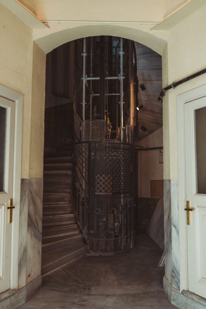 Rustic vintage elevator in historic building hallway with marble accents and open stairway.